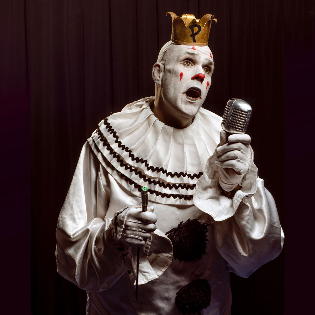 A white-faced clown (Puddles Pity Party) in a ruffled costume and gold crown sings into a vintage microphone against a dark red curtain.