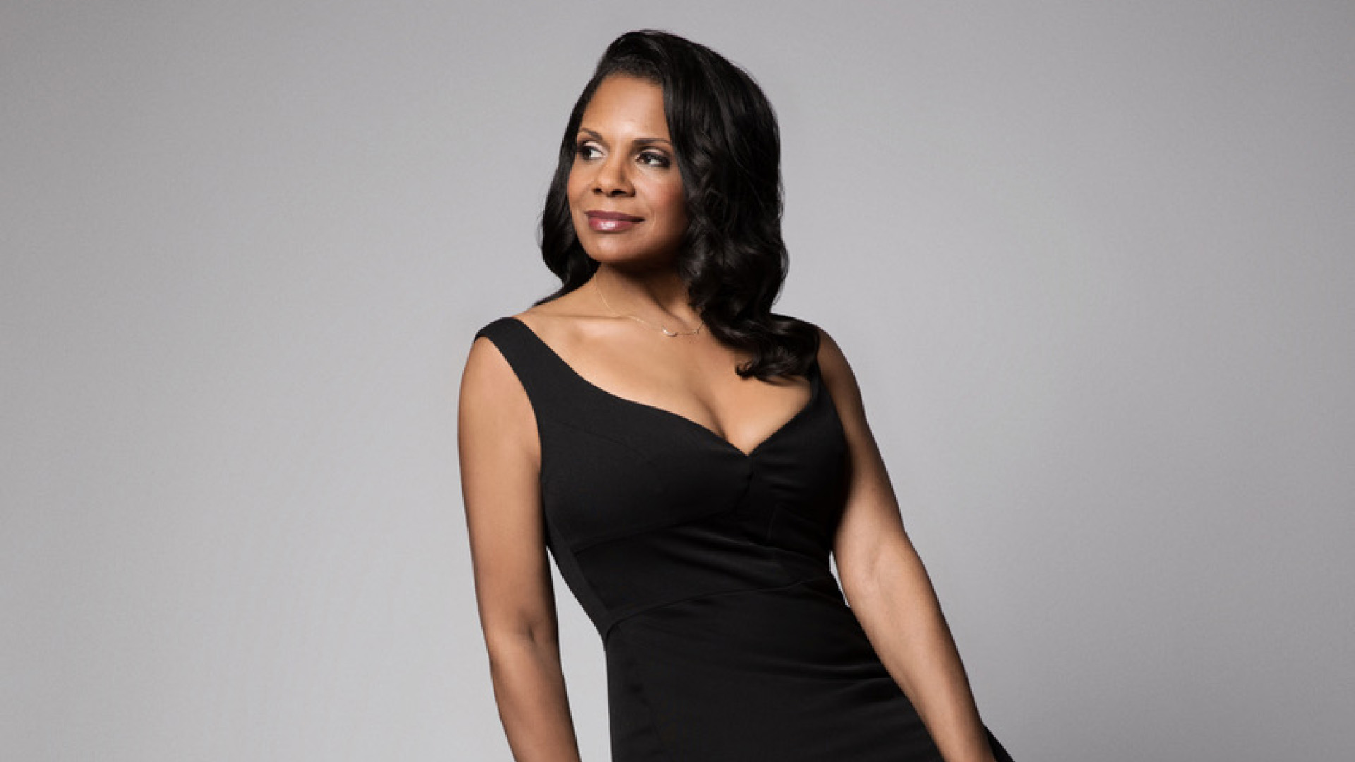 Audra McDonald, in black dress, leaning against stool.