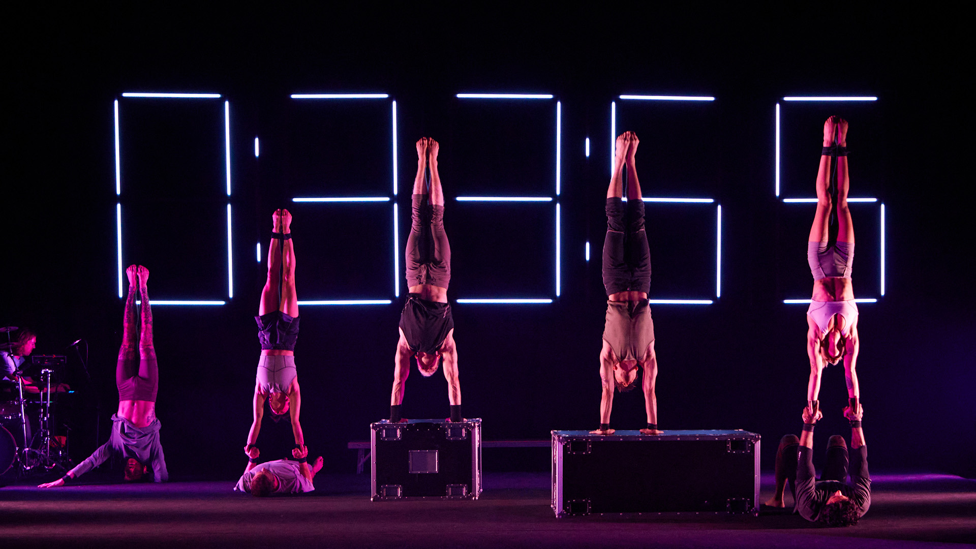 Gravity & Other Myths dancers striking acrobatic poses in front of large digital clock.