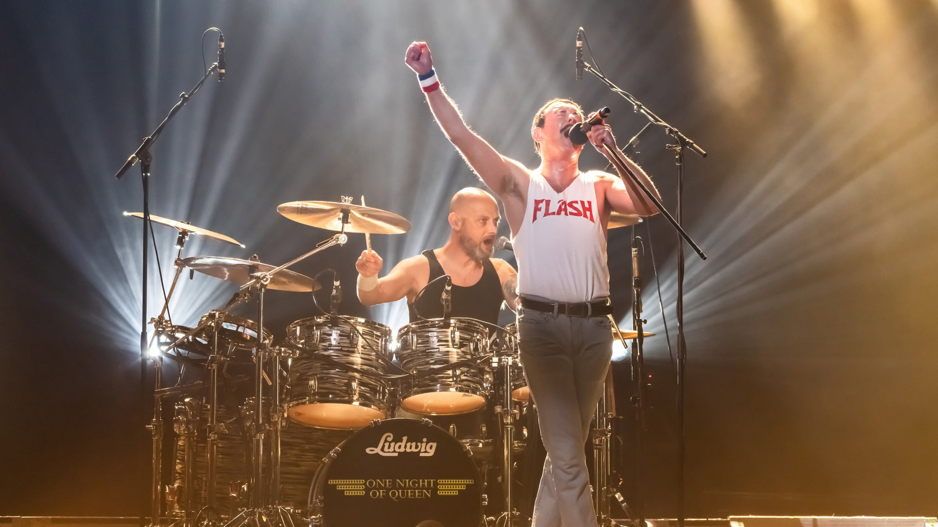Gary Mullen, as Freddie Mercury, singing on stage with fist in air.