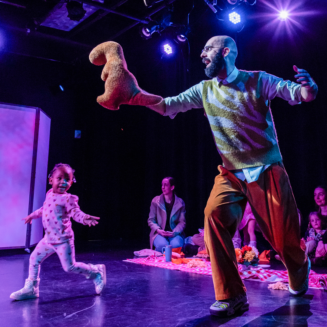 An actor in Dinosaur! holding a small toy dinosaur while a small child stands next to him on stage.